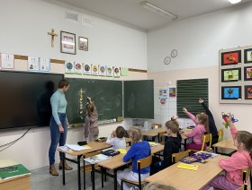 Na zdjęciu widoczna jest sala lekcyjna, a w niej osoba prowadząca zajęcia, dziewczynka, która stoi przy tablicy