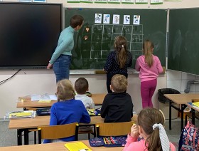 Na zdjęciu widoczna jest sala lekcyjna, a w niej osoba prowadząca zajęcia oraz dziewczynki, które stoją przy tablicy